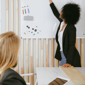Two women engaged in a business presentation with a whiteboard in a modern office setting.